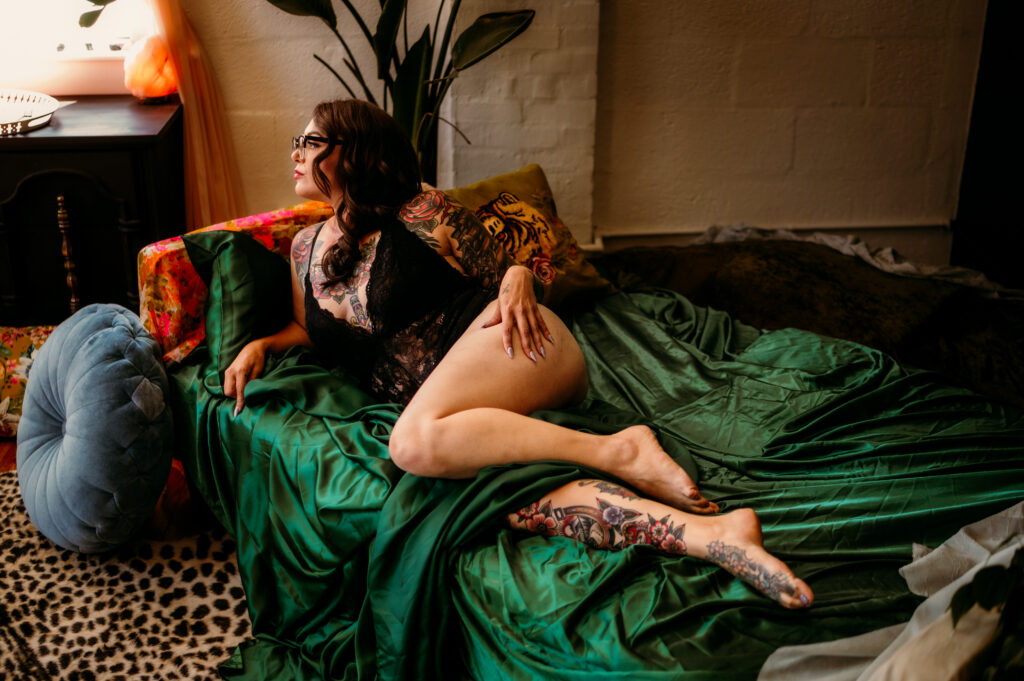 A woman with glasses and tattoos poses in black lingerie on a bed with green satin sheets, surrounded by pillows and soft lighting in a cozy, eclectic Nashville boudoir room.