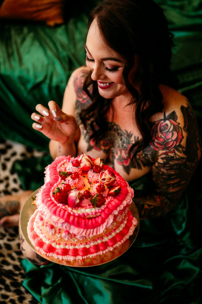 A woman with long dark hair and colorful tattoos smiles while holding a pink, heart-shaped cake decorated with flowers and strawberries. She sits on green silky fabric atop a leopard print rug in a playful Nashville boudoir setting.