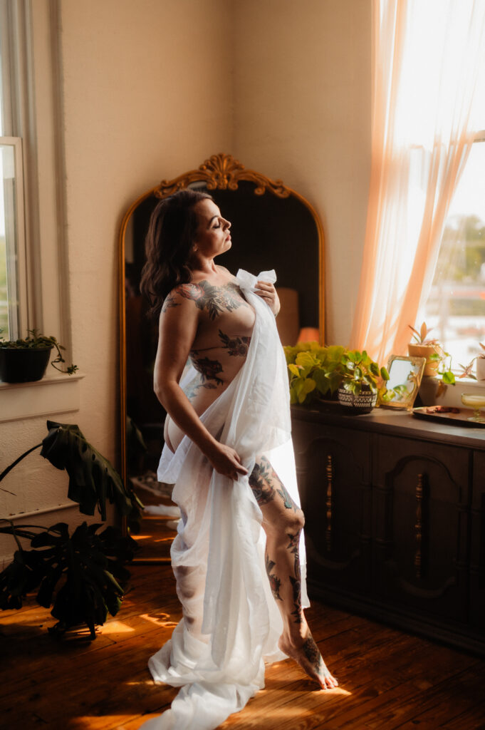 A woman with tattoos stands wrapped in a sheer white sheet near a window, bathed in warm sunlight. In this Nashville boudoir scene, she poses confidently beside a large mirror, surrounded by potted plants and soft, flowing curtains.