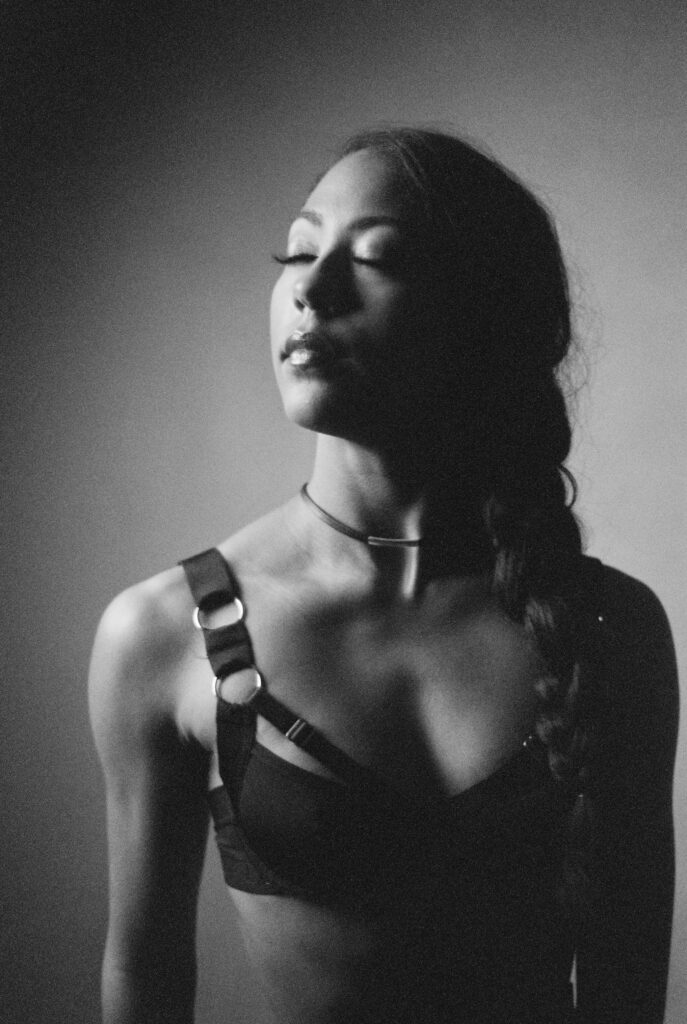 A woman with long braided hair wears a strappy top and choker, standing with her eyes closed and head tilted slightly up, bathed in dramatic, soft lighting against a plain background. This Nashville boudoir photo is made with black and white film.