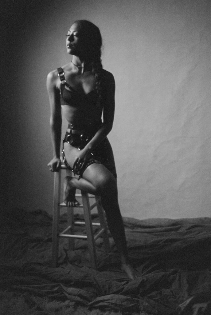 Black and white film photo of a woman in harness-style lingerie sitting sideways on a wooden stool, looking to the left. The setting evokes classic Nashville boudoir style, with fabric-draped background and dramatic shadowed lighting.