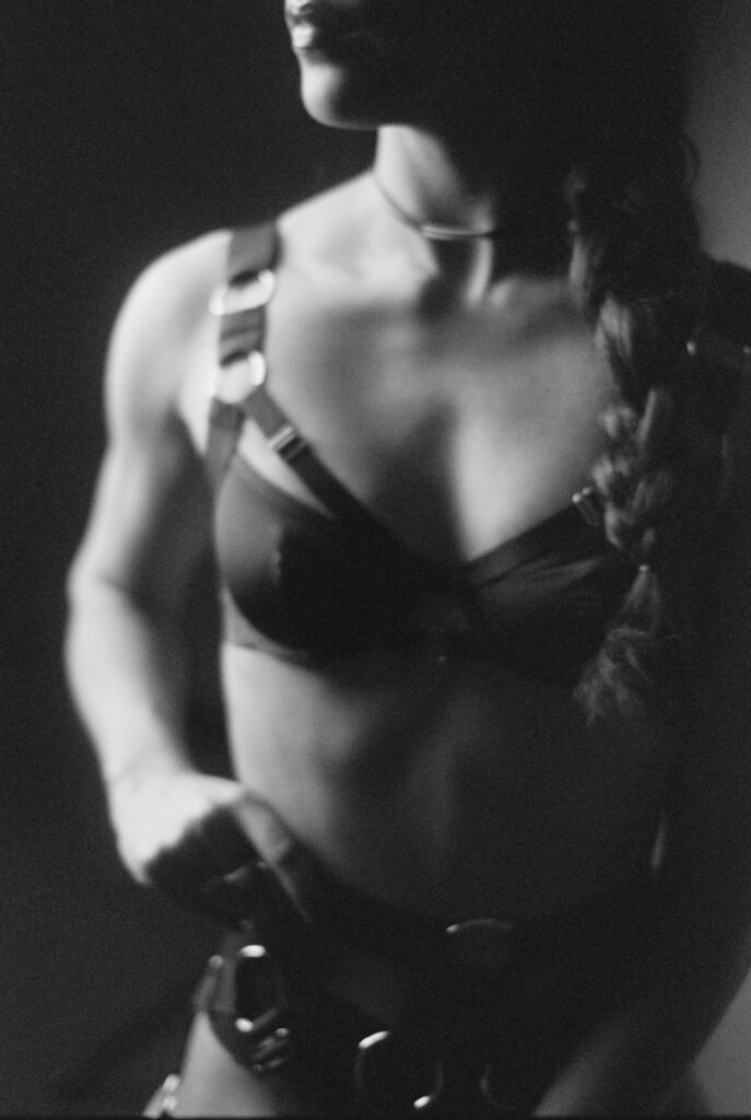 Black and white film photo of a person with a braided hairstyle, wearing a strappy harness and bra. The low lighting highlights their muscular upper body and arm—a bold, artistic Nashville boudoir portrait with their face partially out of frame.