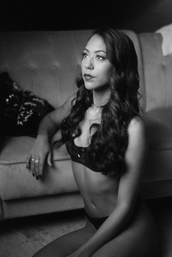 A woman with long wavy hair sits on the floor in front of a sofa, wearing a bra and underwear. She gazes slightly upward with a thoughtful expression in this black and white film Nashville boudoir image.