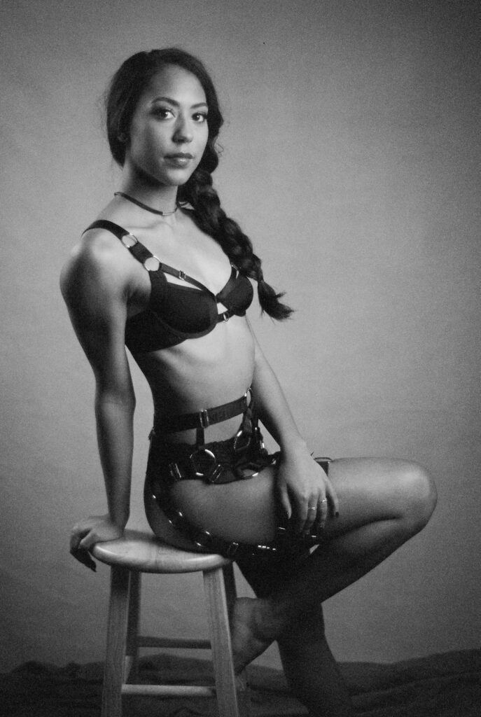 A woman in lingerie and harness sits sideways on a wooden stool, facing the camera in a dimly lit, black-and-white film studio portrait, capturing the sultry elegance of a Nashville boudoir session.
