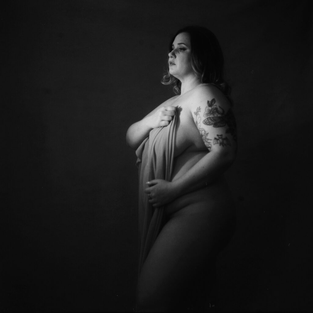 A black-and-white photo of a woman standing, partially draped in fabric, covering her body. She has a tattoo on her upper arm and gazes pensively into the distance—a striking example of Nashville boudoir style against a dark background.