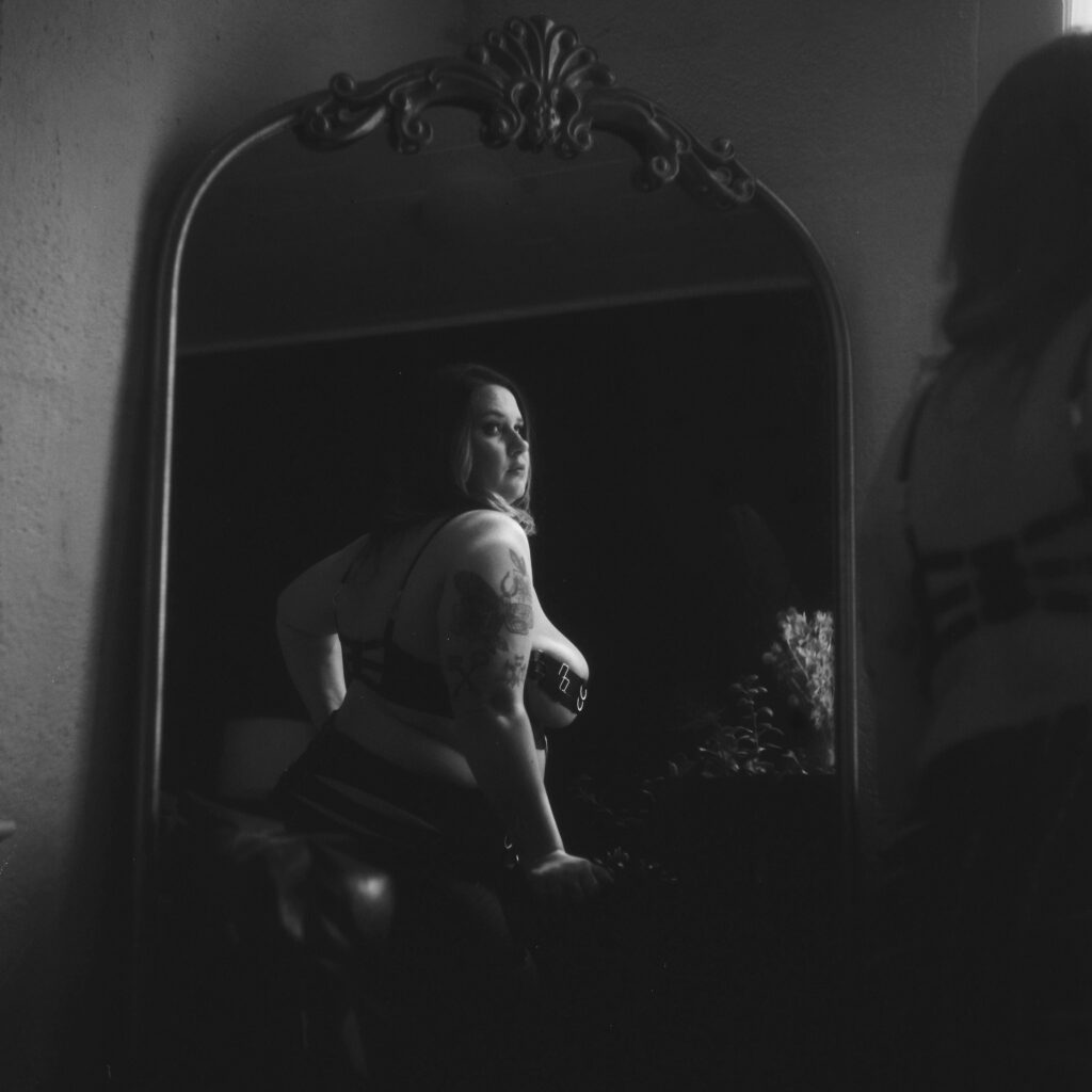A woman in lingerie poses confidently before an ornate mirror, her tattooed arm visible, with soft, dramatic lighting creating a moody, intimate black-and-white Nashville boudoir atmosphere.