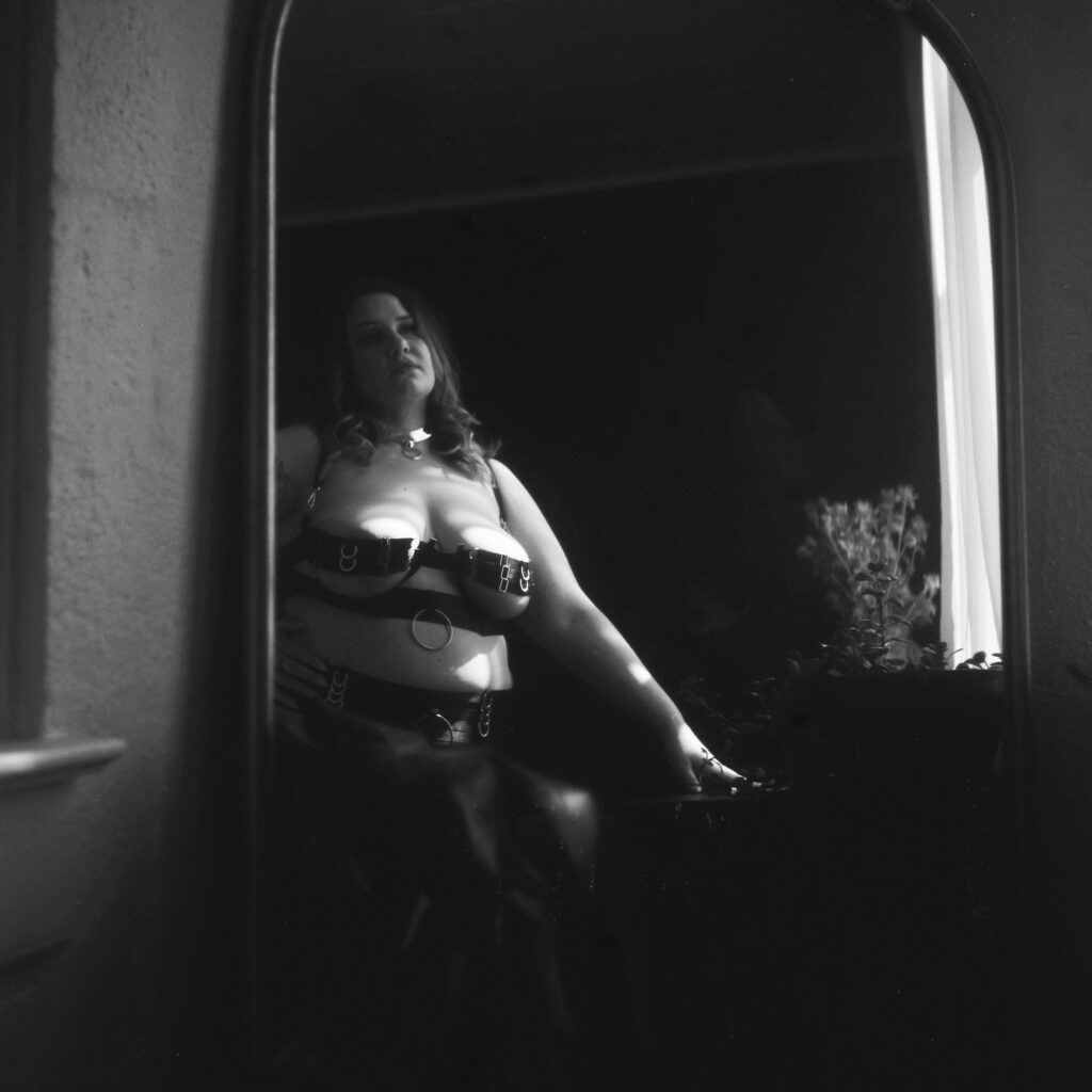 A woman dressed in black leather harness lingerie is seen through a mirror, posing confidently with a hand on her hip in a dimly lit Nashville boudoir, soft window light accentuating her silhouette.