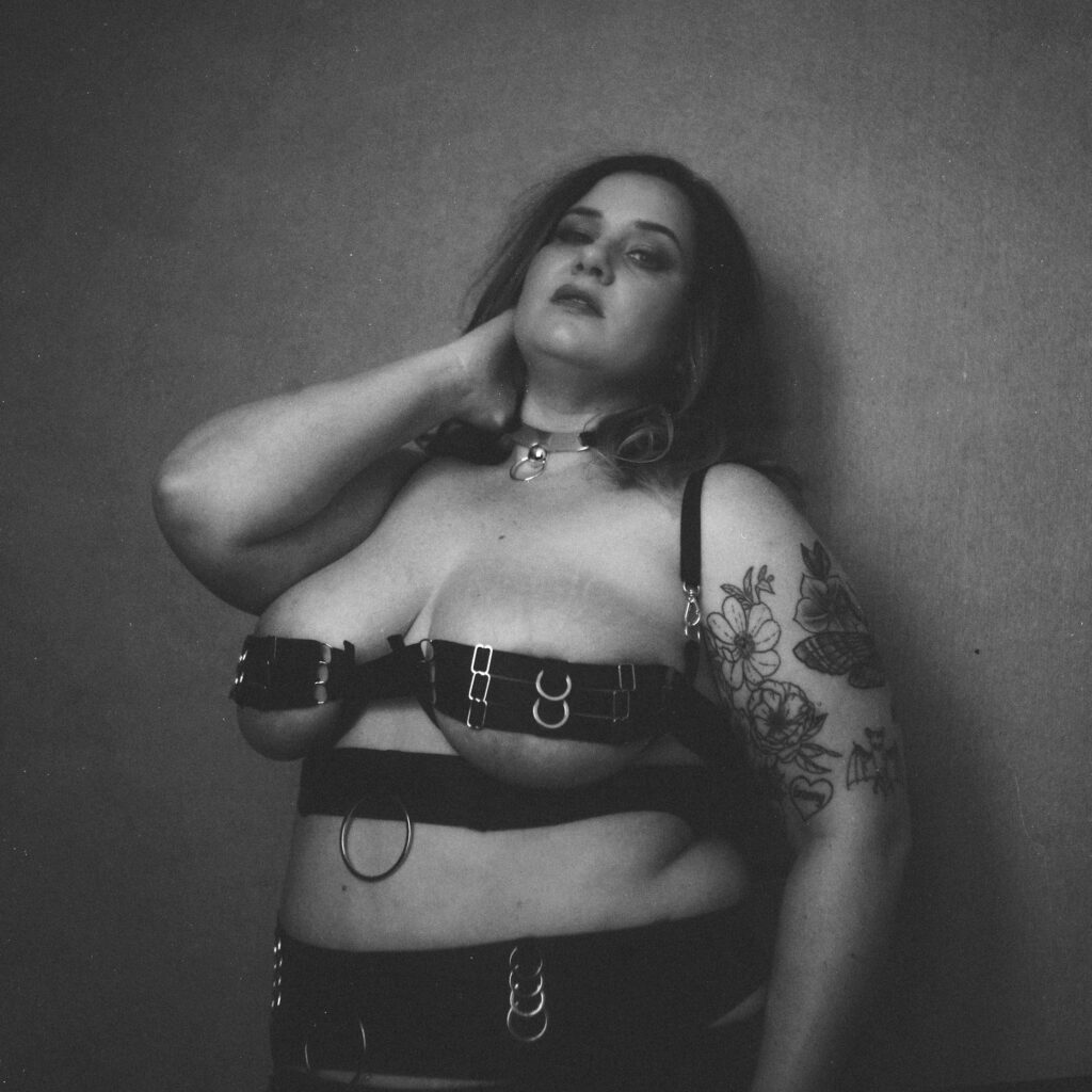 A woman with long hair poses confidently in a strappy black harness outfit, wearing a choker necklace. She has a floral tattoo on her arm and stands against a plain background, capturing the bold spirit of Nashville boudoir in black and white.