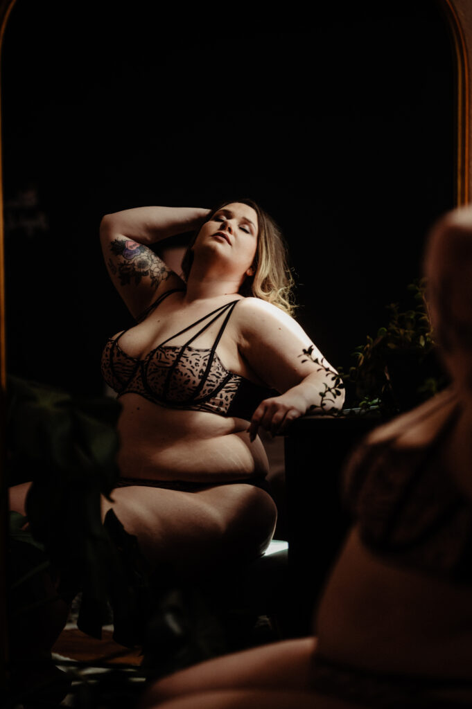 A person with long hair and tattoos poses confidently in lingerie, sitting in front of a mirror with plants nearby. Dramatic, low lighting enhances this Nashville boudoir scene.