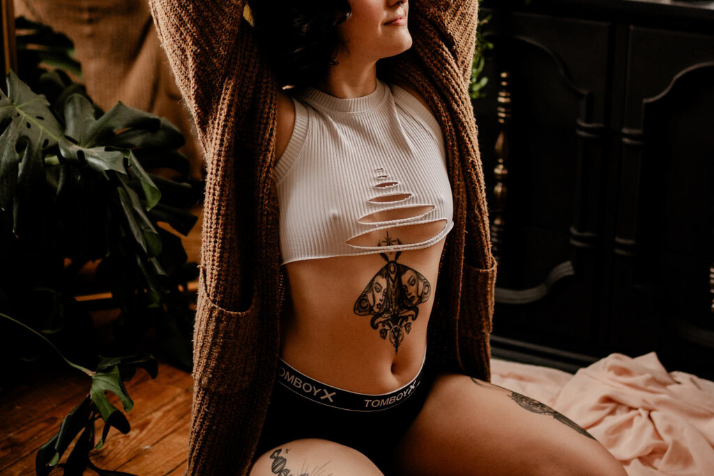 A person sits on the floor indoors, wearing a ripped white crop top, black underwear, and a brown cardigan with arms raised. Visible tattoos decorate their torso and thigh. Nashville boudoir style is reflected in the houseplants and chic furniture in the background.
