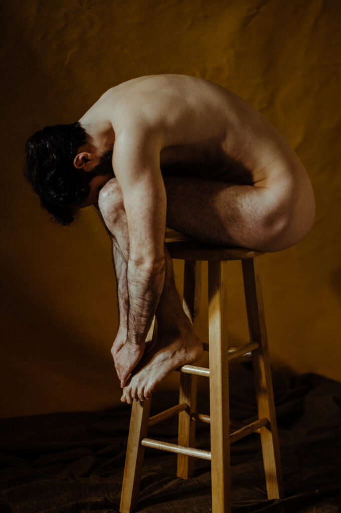A nude person sits curled up on a wooden stool against a brown background, arms hugging their legs and head resting on their knees, capturing a contemplative and introspective pose in this Nashville boudoir-inspired scene.