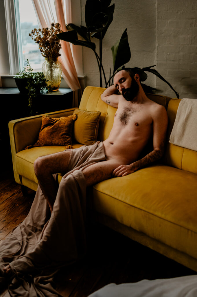 A bearded man is reclining nude on a yellow sofa, partially covered by a beige sheet, with plants and soft sunlight in the background, embodying the relaxed and intimate vibe of a Nashville boudoir.