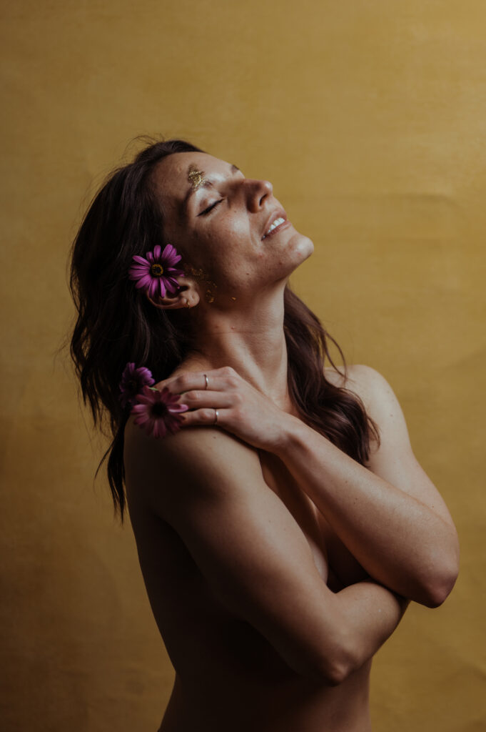 A woman with closed eyes smiles softly against a yellow background, holding pink flowers near her shoulder and ear. Gold flakes accent her face and shoulder as she poses in a gentle, nashville boudoir-inspired style, arms crossed delicately over her chest.