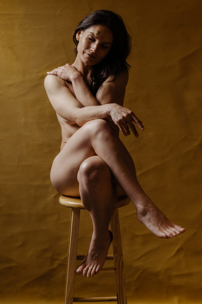 A nude woman with long dark hair sits cross-legged on a wooden stool against a brown background in this Nashville boudoir scene, covering her chest with one arm and resting her other hand on her knee, looking downward with a gentle expression.