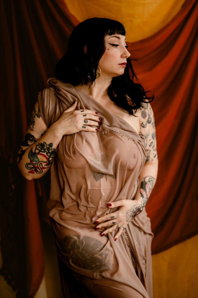 A woman with dark hair and tattoos poses in front of draped brown and gold fabric, wearing a sheer, flowing beige gown. Captured in a Nashville boudoir style, she rests one hand on her chest and the other on her waist, gazing to the side.