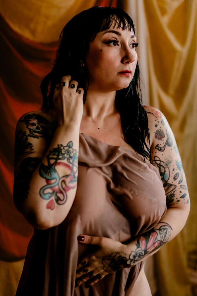 A woman with dark hair and tattoos on her arms and hands stands against a yellow draped background, covering her chest with sheer brown fabric. Capturing a thoughtful gaze, this Nashville boudoir moment is both intimate and striking.