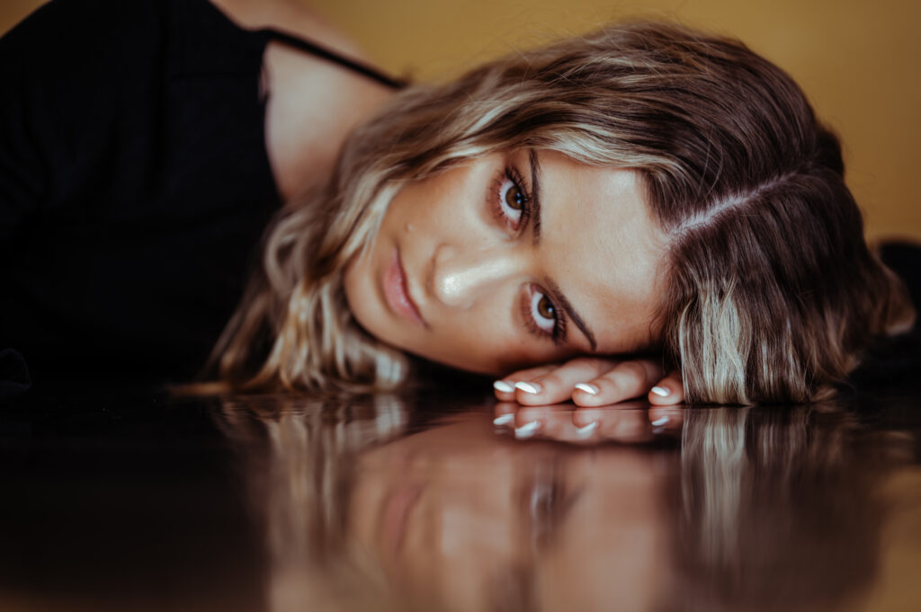 A woman with wavy blonde hair lies on her side, resting her head on her hand against a reflective surface, gazing directly at the camera in this intimate Nashville boudoir scene. The background is blurred with warm tones.