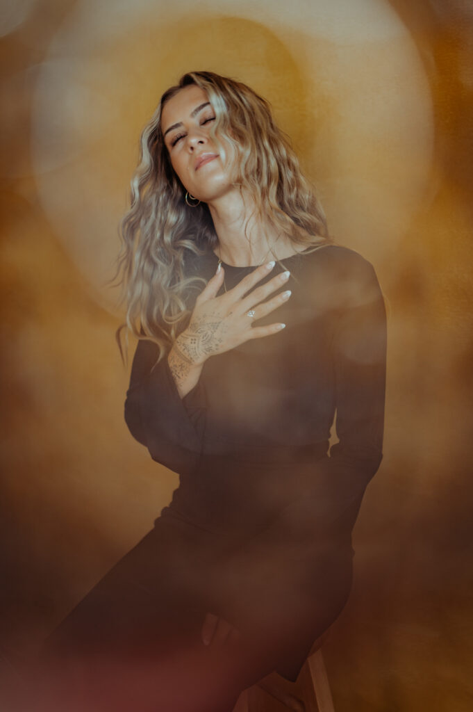 A woman with long wavy hair wearing a black dress sits on a stool against a warm, brown background. In this Nashville boudoir scene, she touches her chest with one hand, eyes closed, appearing serene and thoughtful.