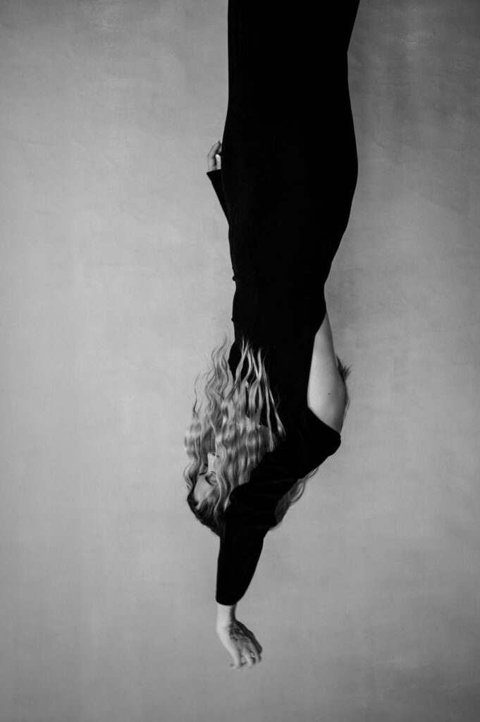A person with wavy hair, dressed in black, hangs upside down against a plain background. Their arm is extended downward and their face is hidden. This striking black and white image evokes a moody Nashville boudoir aesthetic.