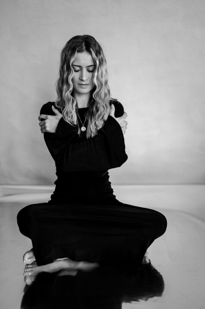 A woman with long wavy hair sits cross-legged on the floor, eyes closed, hugging herself in a tender embrace. She wears a long black dress and a necklace. This black-and-white image captures intimate Nashville boudoir vibes.