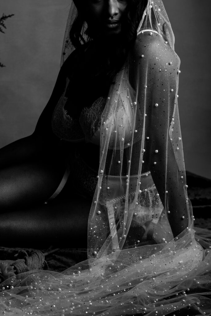 A woman in lacy lingerie poses while sitting, draped in a sheer, pearl-studded veil. The black and white photo captures the texture and shimmer, creating a dramatic, elegant mood perfect for a Nashville bridal boudoir session.