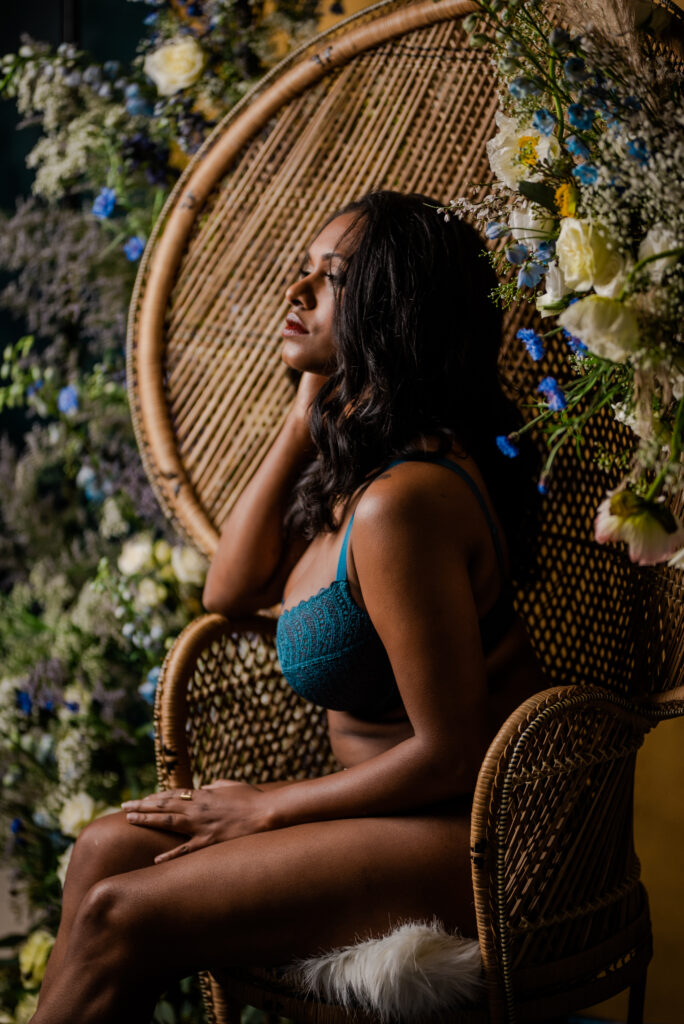 A woman in teal lingerie sits on a wicker chair surrounded by flowers, gazing thoughtfully to the side as soft light highlights her face and hair in this elegant Nashville boudoir scene.