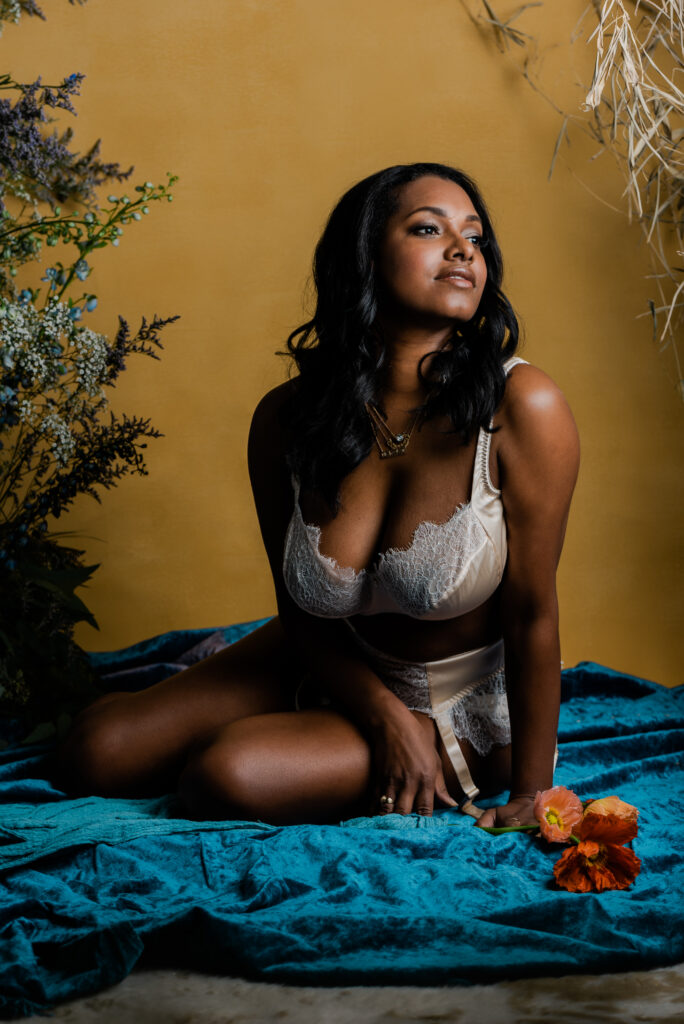 A woman in cream-colored lingerie sits on teal velvet fabric with flowers around her, looking off to the side. The warm yellow background and lush plants create a dreamy Nashville bridal boudoir vibe.