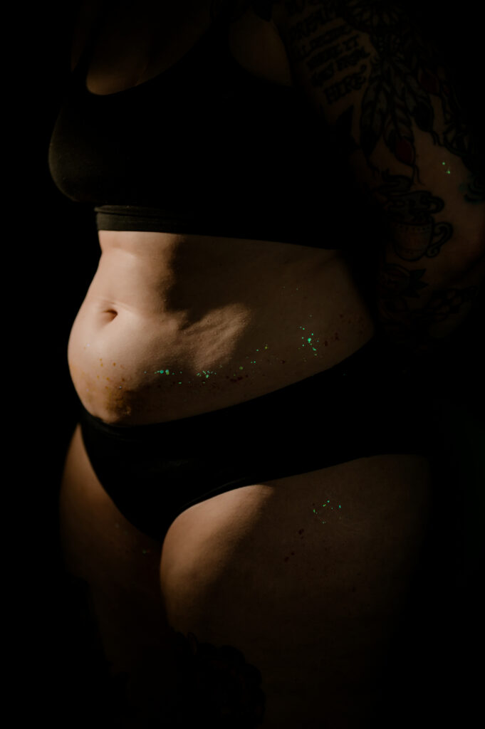A person wearing black underwear and a black bra stands in partial shadow, glitter on their stomach and hip catching the light. Tattoos are visible on their arm, while the lighting, perfect for a Nashville boudoir session, highlights the natural curves of their body.
