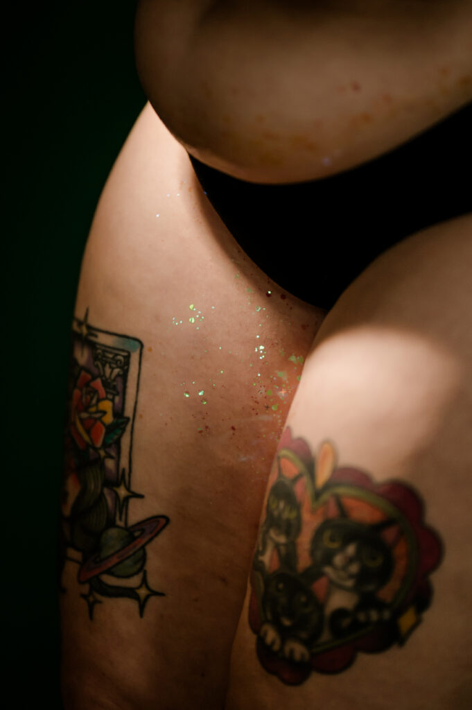 A close-up of a person's upper thighs and lower torso, wearing black underwear. Their skin sparkles under the light, with two colorful tattoos: a flower in a frame and three cats in a heart-shaped border—capturing the spirit of Nashville boudoir.