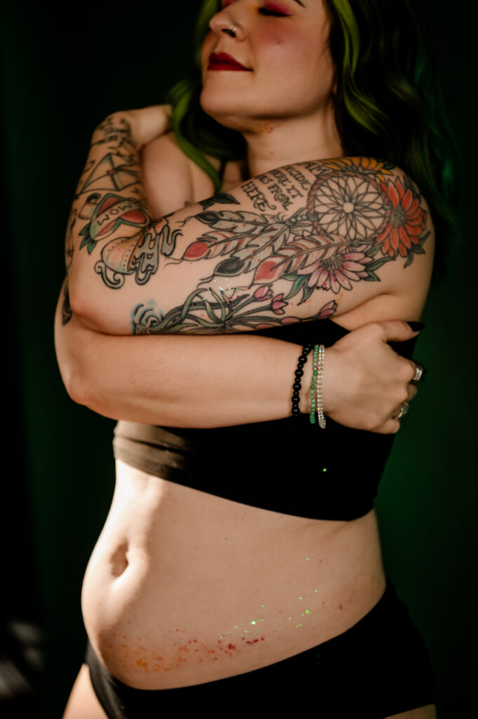 A person with colorful tattoos smiles and hugs themselves, wearing a black bra and underwear in a Nashville boudoir. Their eyes are closed, showing self-acceptance. Glitter decorates their belly, and a bracelet is visible on their wrist.