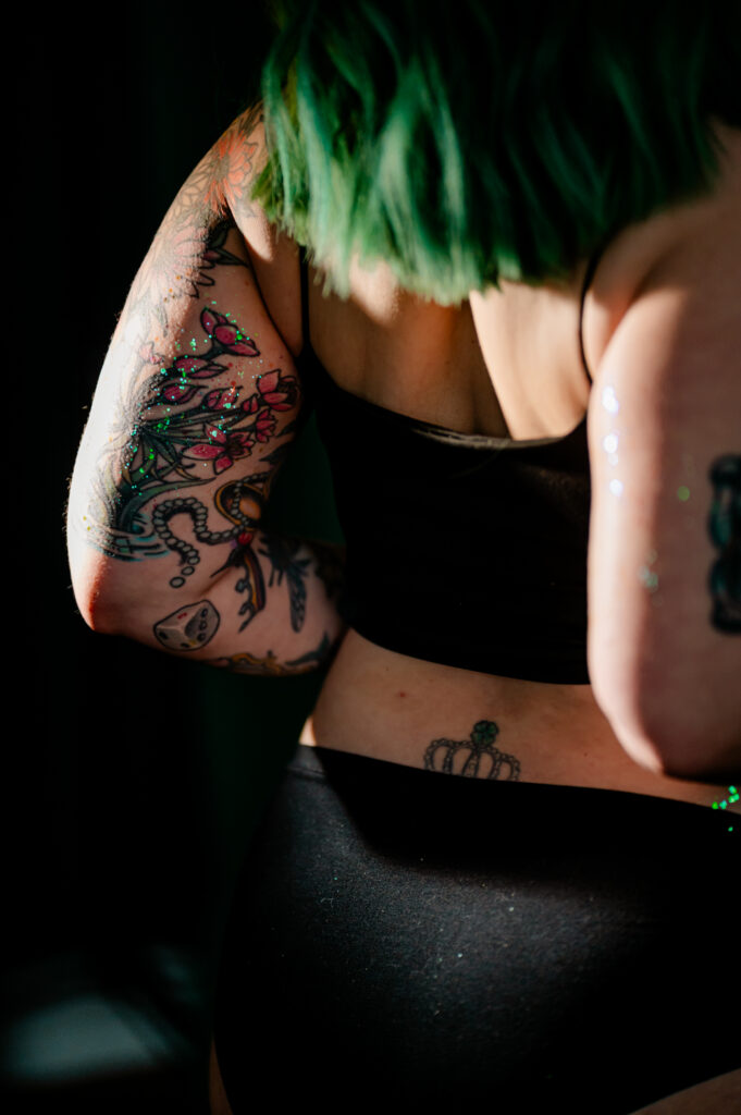 A person with green hair wearing black underwear and a black top, showing tattoos on their arms and lower back, stands in soft lighting, capturing a tasteful Nashville boudoir vibe.