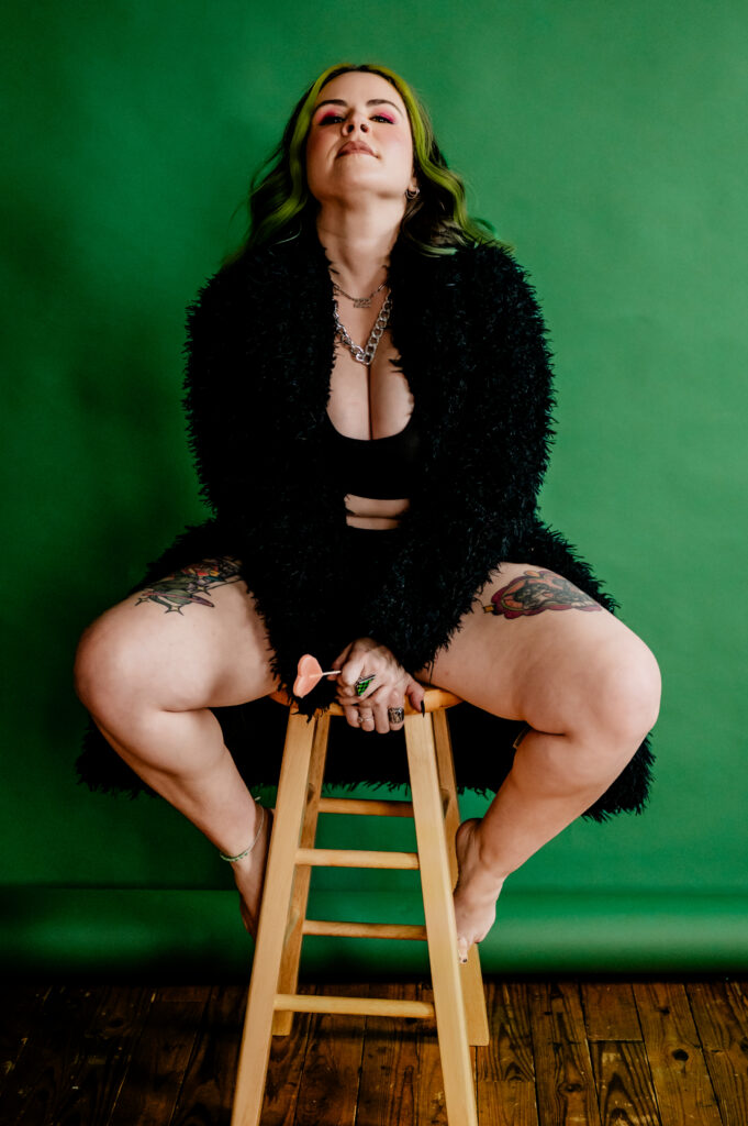 A person with green hair and pink eyeshadow sits on a wooden stool against a green background, wearing a black fuzzy coat and black top, displaying tattoos on their thighs and exuding Nashville boudoir confidence as they look upward.
