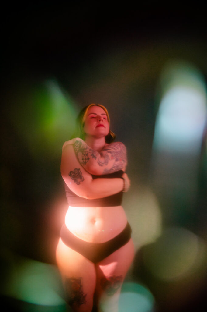 A woman with tattoos, wearing black underwear, stands with her arms wrapped around herself and eyes closed against a softly blurred background with colorful light effects, capturing the intimate vibe of a Nashville boudoir session.