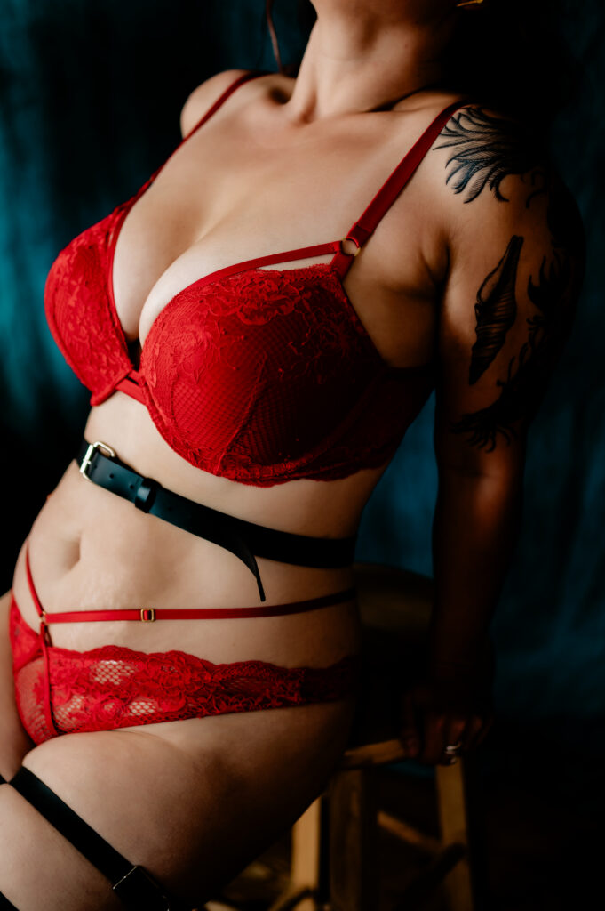 A woman wearing red lace lingerie poses while sitting on a wooden stool in this Nashville boudoir scene. Her head is slightly tilted back, revealing a black tattoo on her upper arm and shoulder against a dark, blurred background.