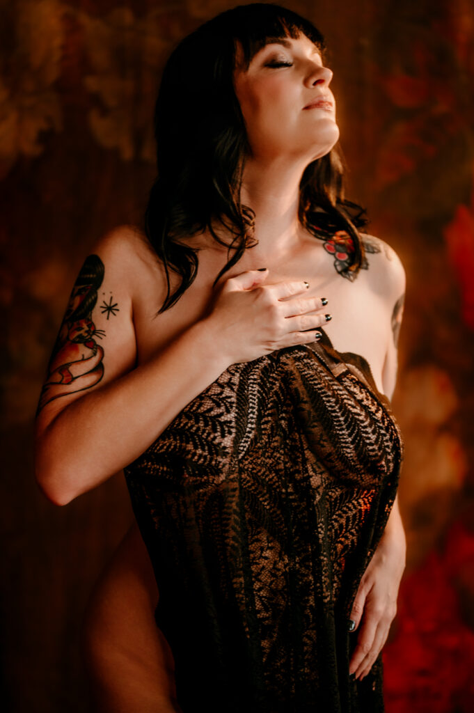 A woman with dark hair and tattoos stands against a warm, textured background, eyes closed, gently holding a black lace fabric over her chest and abdomen, exuding the serene confidence of a Nashville boudoir session.