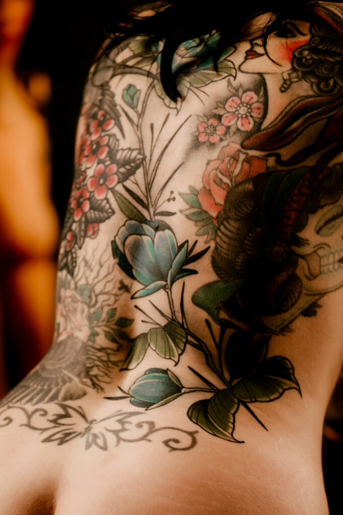 A person's bare back covered in colorful tattoos, featuring flowers, leaves, and intricate designs. The image evokes a nashville boudoir vibe, focusing on detailed artwork and highlighting vibrant reds, greens, and blues.