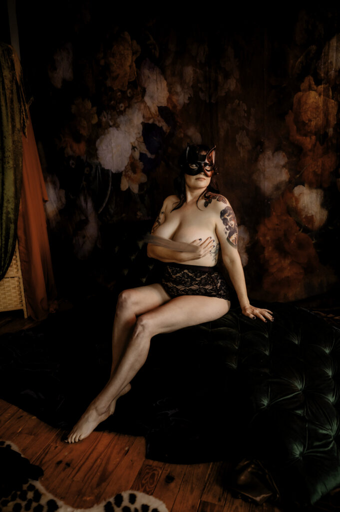 A person wearing a decorative mask and black lace lingerie poses on a green tufted ottoman in a dimly lit room with a floral backdrop and wooden floor, showcasing visible tattoos—a striking moment in this Nashville boudoir setting.