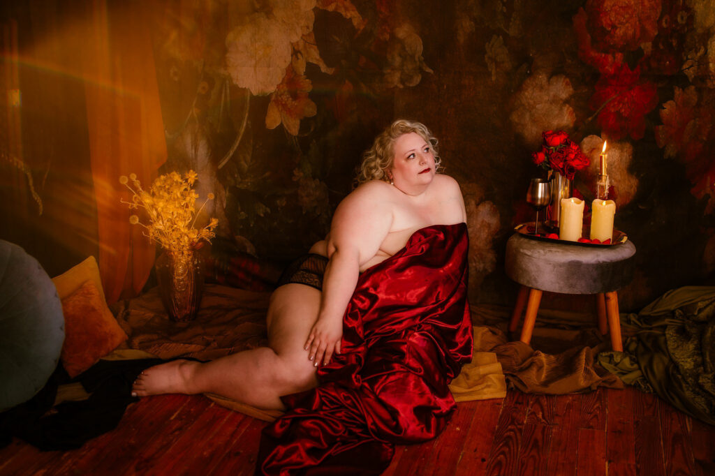 A person draped in red satin poses seated on the floor against a dramatic, floral-patterned backdrop—capturing the intimate elegance of a Nashville boudoir—surrounded by candles, flowers, and wine, all bathed in warm, golden light.