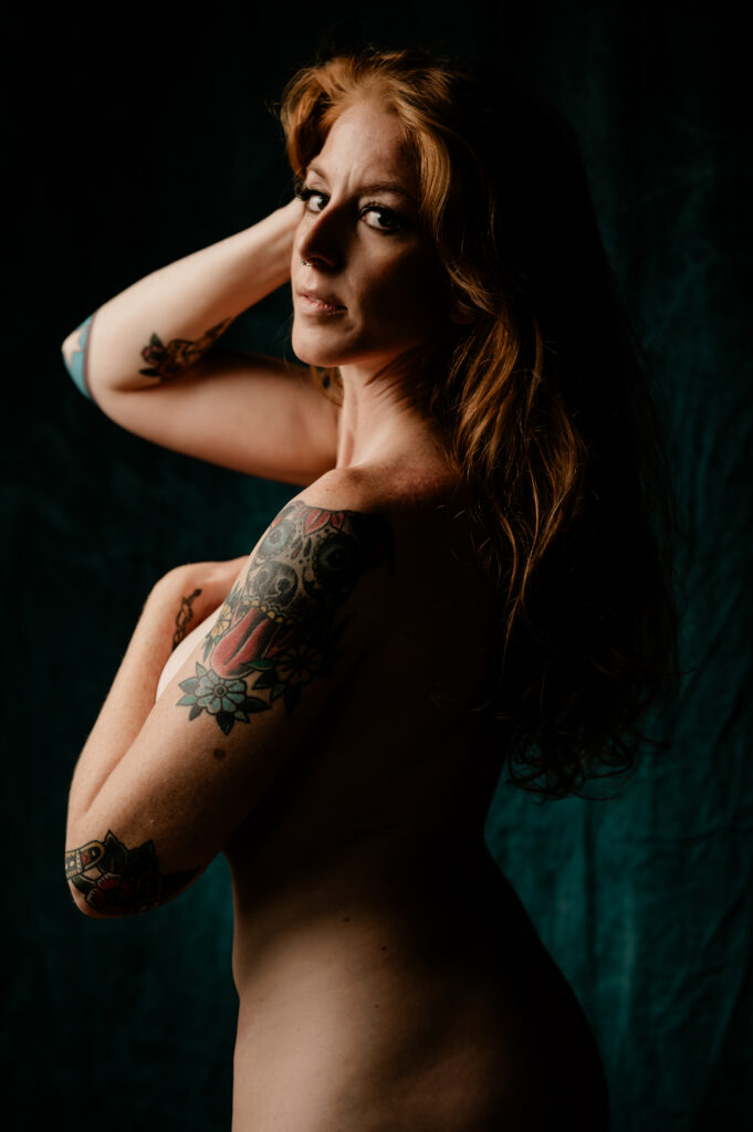 A nude woman with long red hair poses against a dark background in a Nashville boudoir style, her arm covering her chest and showcasing colorful tattoos on her shoulder and arm as she looks over her shoulder toward the camera.