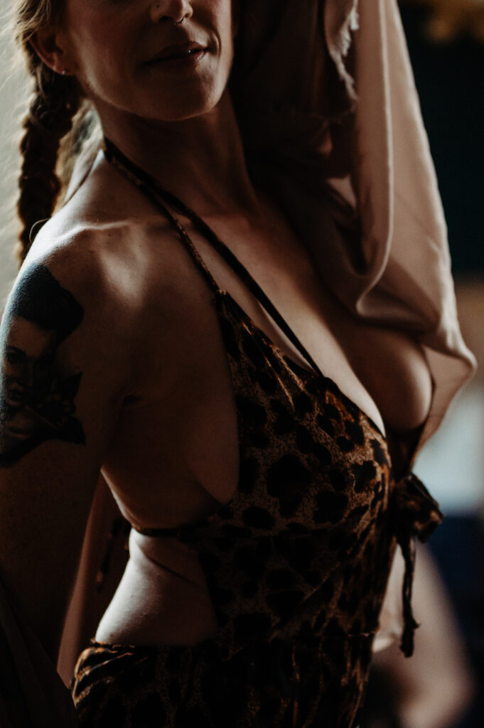 A woman with braided hair wears a leopard-print dress, partially covered by a draped fabric. She has a tattoo on her upper arm and poses in a dimly lit Nashville boudoir setting.