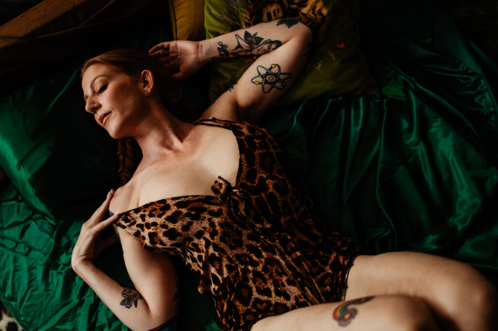 A woman in a leopard print outfit reclines on green satin sheets, eyes closed and arms stretched above her head, displaying various tattoos—a stunning example of Nashville boudoir artistry.