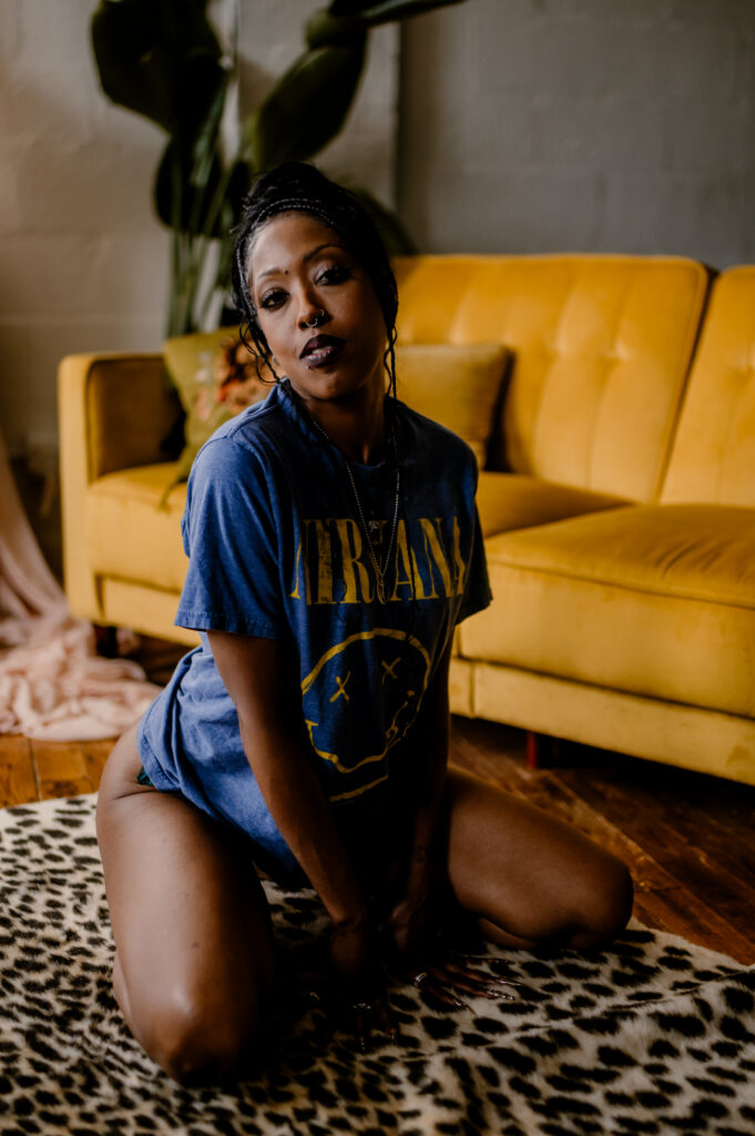 A person wearing a blue Nirvana T-shirt kneels on a leopard-print rug in front of a yellow couch, with green plants and soft natural light in the background, capturing the relaxed vibe of a Nashville boudoir.