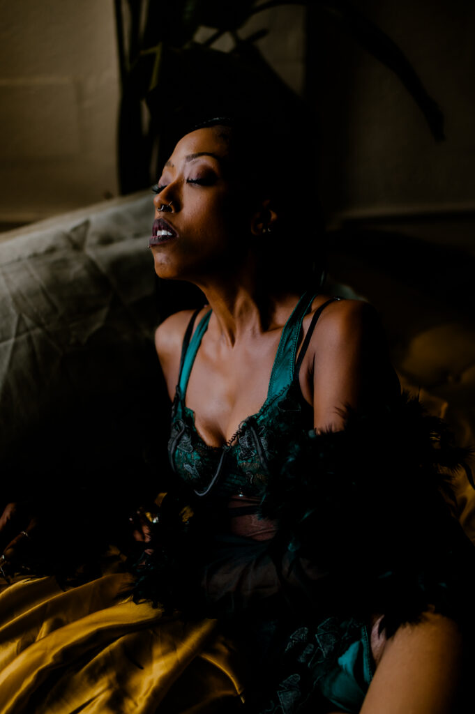 A woman in green lingerie and a feathered garment sits on a bed, eyes closed, illuminated by soft, moody lighting—a captivating scene from a Nashville boudoir session.
