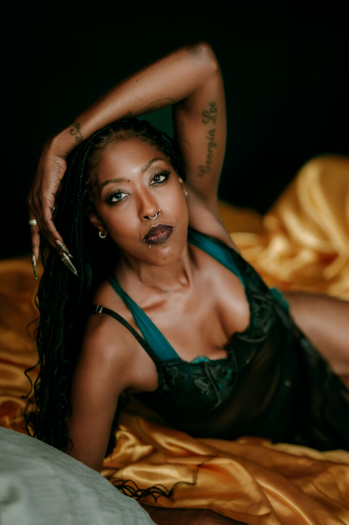 A woman with long braided hair and tattoos poses confidently on a bed with yellow sheets, wearing dark lingerie. This Nashville boudoir scene captures her piercing, intense expression as she looks directly at the camera.