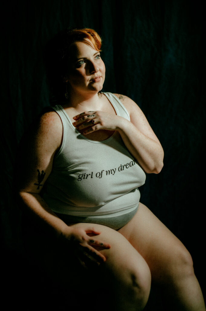 A woman with fair skin and short auburn hair sits against a dark backdrop, exuding Nashville boudoir elegance in a light tank top that says Girl of my dreams. She gazes upward with one hand on her chest and the other resting on her leg.