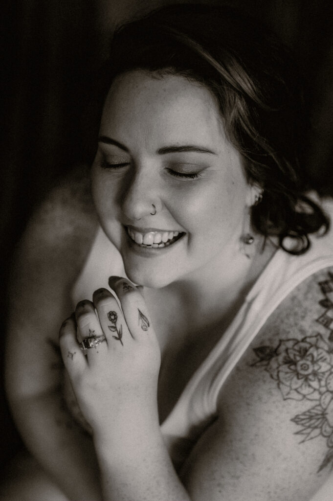 A woman with tattoos on her arm and fingers smiles with her eyes closed, resting her hands near her face. She wears a nose ring, earrings, and a sleeveless top in this black and white Nashville boudoir portrait.