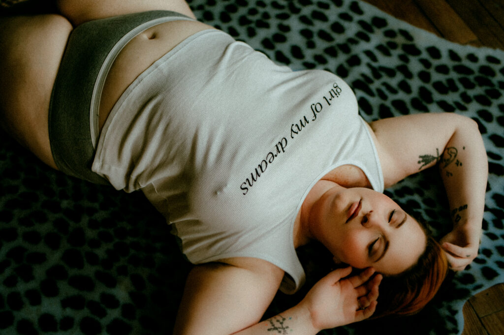 A person with light skin and short hair lies on a patterned blanket, wearing a white tank top that reads girl of my dreams and gray underwear. Showcasing tattoos on their arms, they appear relaxed with eyes closed in this Nashville boudoir scene.