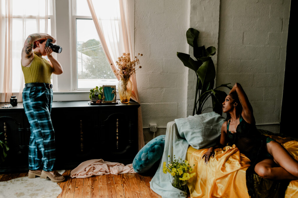 A photographer in a yellow top and blue plaid pants takes a picture of a woman posing on a bed with yellow sheets, capturing a Nashville boudoir vibe in a sunlit room filled with plants and large windows.