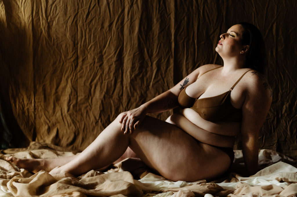 A woman in brown lingerie sits on a fabric-draped floor with legs extended, eyes closed and head tilted upward, bathed in warm natural light—a moment of tranquil self-perception against a textured brown backdrop.