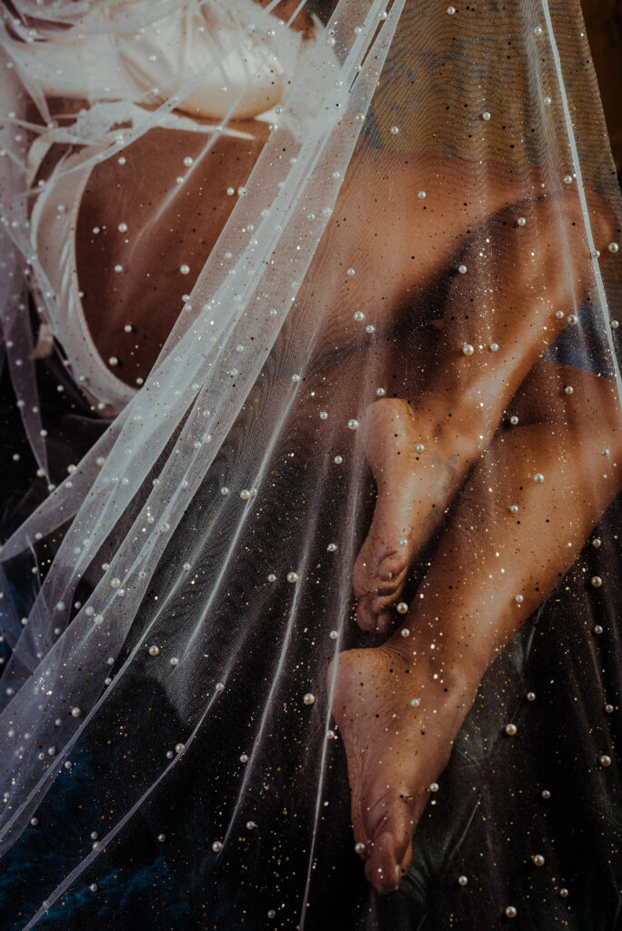 A person wearing white lingerie sits with legs crossed, partially covered by a sheer, pearl-embellished fabric. The bridal boudoir lighting highlights the smooth texture of their skin beneath the delicate material.